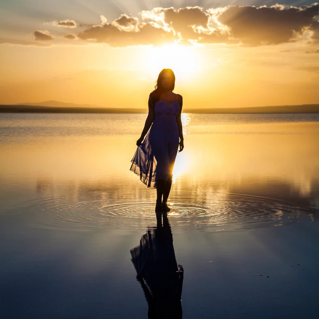 Une femme en robe légère marche dans l'eau peu profonde au coucher du soleil, créant des ondulations autour de ses pieds. Le soleil bas illumine la scène avec des tons dorés, projetant une silhouette et un reflet délicat sur la surface calme de l'eau. Le ciel, parsemé de nuages, ajoute une touche de sérénité et de poésie.