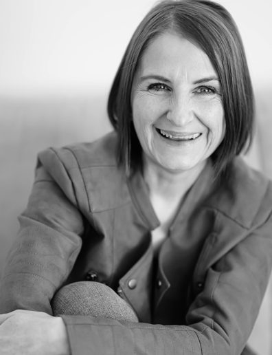 Portrait en noir et blanc de Virginie Petitjean, souriante, assise dans une posture détendue.