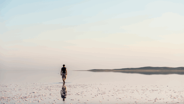 Personne marchant seule sur une étendue d’eau peu profonde, dont la surface miroitante reflète le ciel pastel et l’horizon lointain.